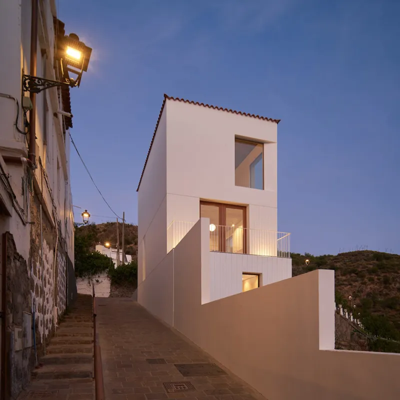 White cubic house wedged into a narrow hillside street at dusk with lit windows