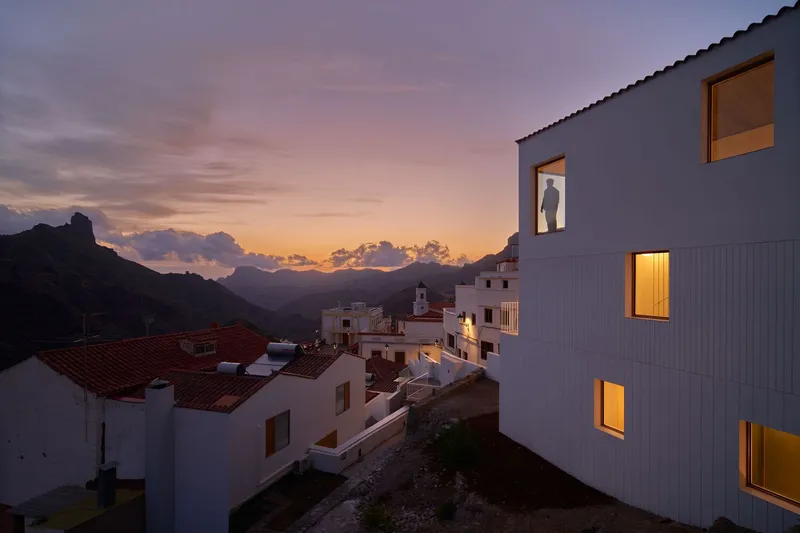 White corrugated facade with illuminated windows rising above a hillside village at dusk