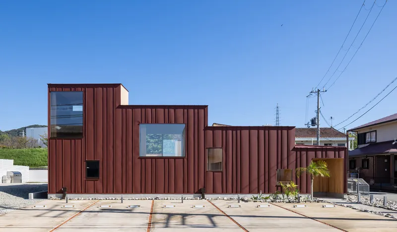 Street elevation with vertical rust-red metal cladding, stepping volumes, and gravel forecourt under power lines