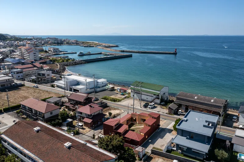 Aerial view of low-rise red metal-roofed buildings along a coastal road with harbor breakwater and sea beyond