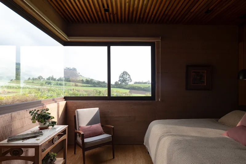 Bedroom with rammed earth walls, slatted timber ceiling, and horizontal window overlooking farmland and hills