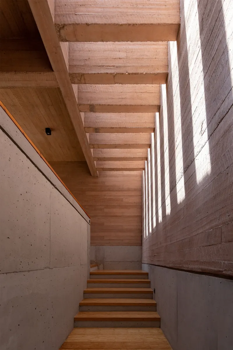 Timber staircase rising between concrete walls with slotted clerestory casting striped shadows down the steps