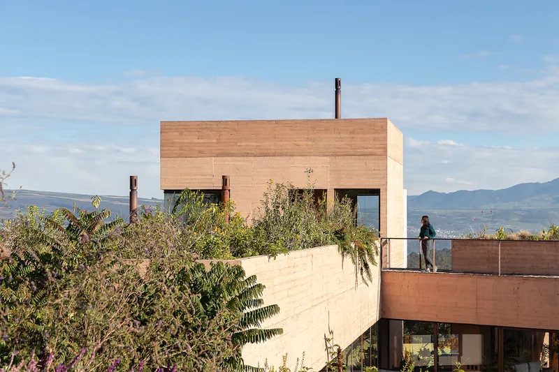 Planted roof terrace with person standing beside the upper rammed earth volume and valley beyond