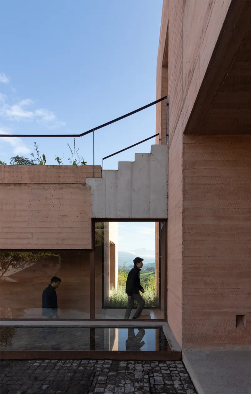 Exterior stair climbing past rammed earth walls with figure walking through glazed corridor below