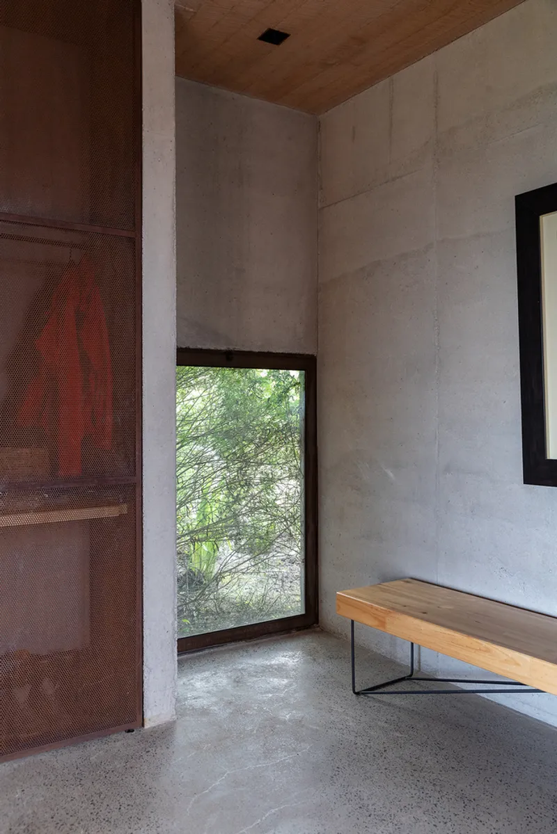 Entry corner with perforated metal screen, concrete walls, timber bench, and horizontal window framing garden foliage