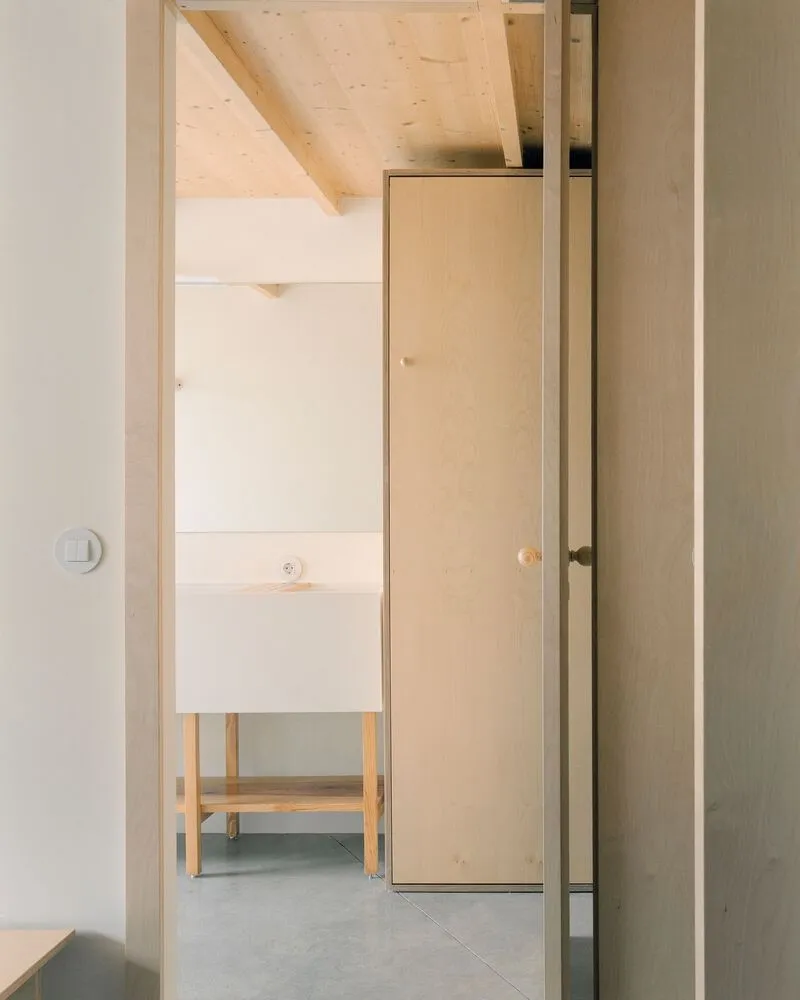 Hallway view through plywood doorway toward freestanding sink beneath sloped timber ceiling