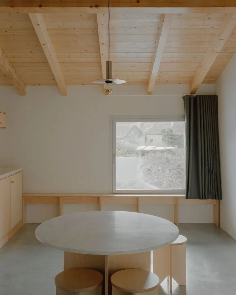 Dining area with round concrete table and exposed timber rafters framing a window view of snowfall