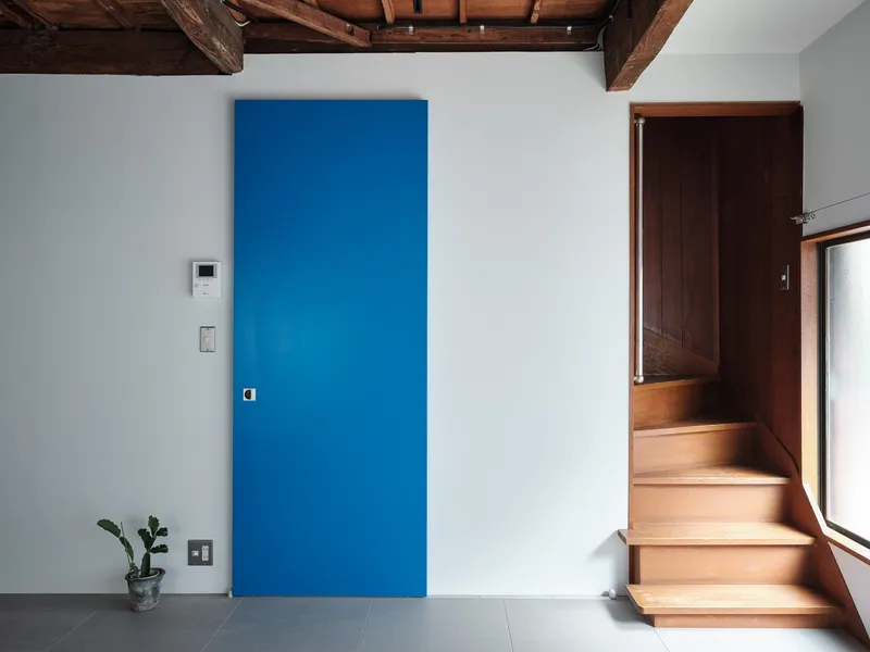 Blue door panel and timber staircase rising along white wall beneath exposed wooden beams with small plant