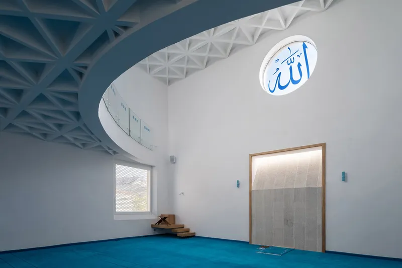 Curved prayer hall wall with geometric ceiling panels and a perforated window casting patterned shadows