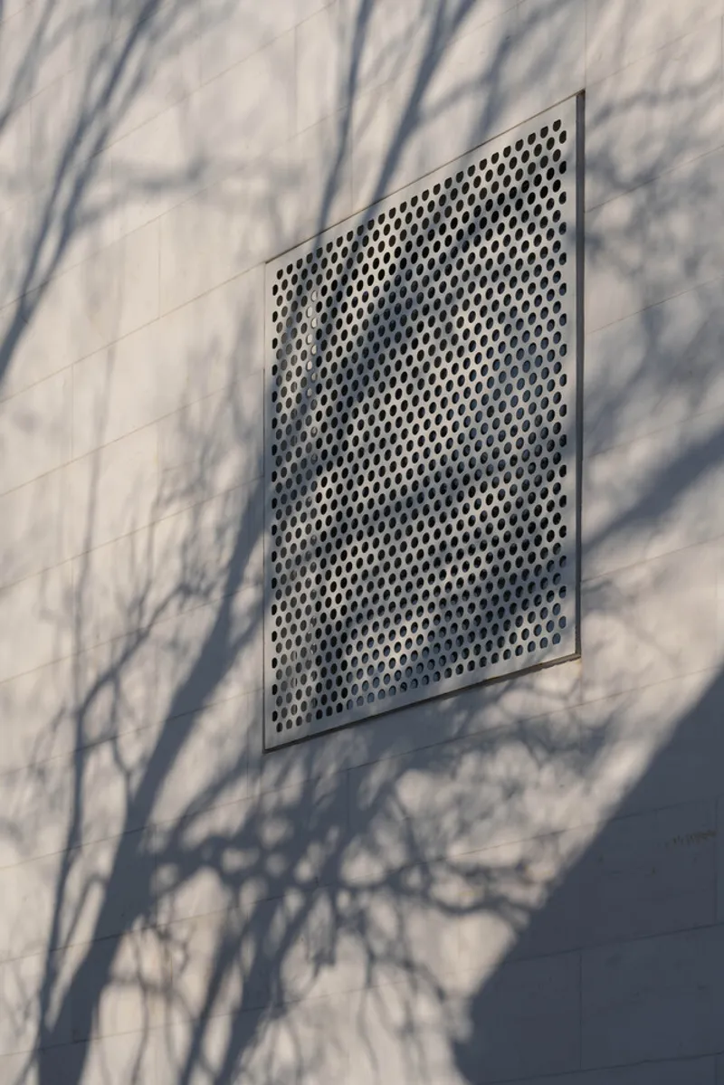 Perforated metal panel set into white stone wall with tree branch shadows cast across the surface