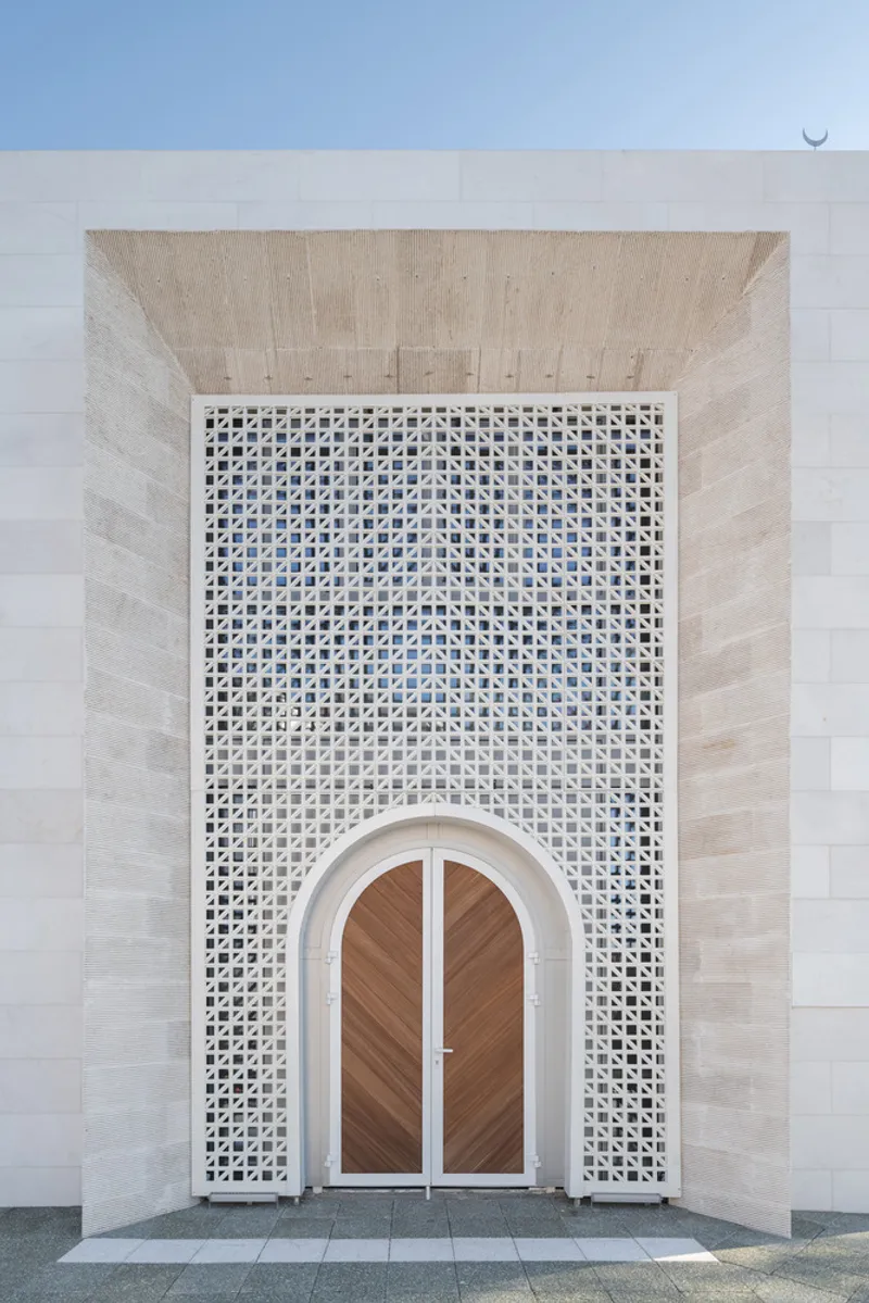 Entrance with timber arched doors framed by perforated white screen under recessed stone canopy