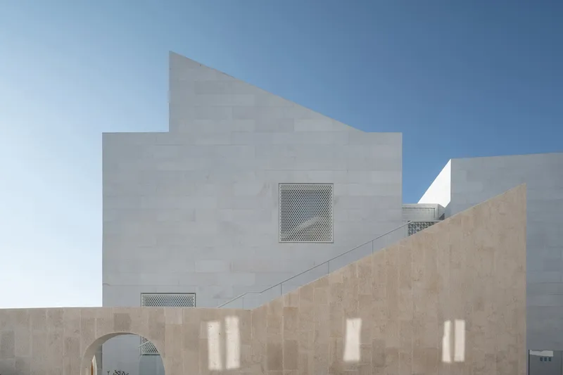 White plastered wall with perforated screens above stone wall casting shadow patterns in daylight