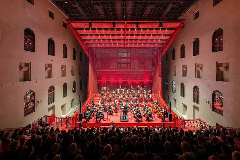 View across the auditorium showing tiered seating and orchestra performing under red theatrical lighting at night