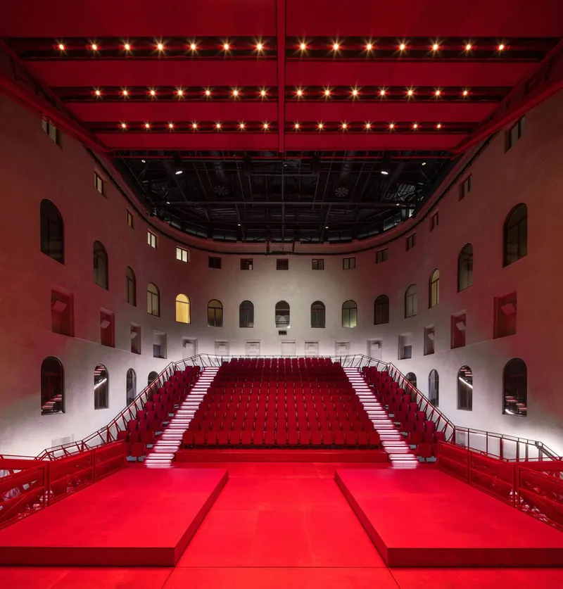 Stage view showing red stepped seating platforms flanked by arched window openings in plaster walls