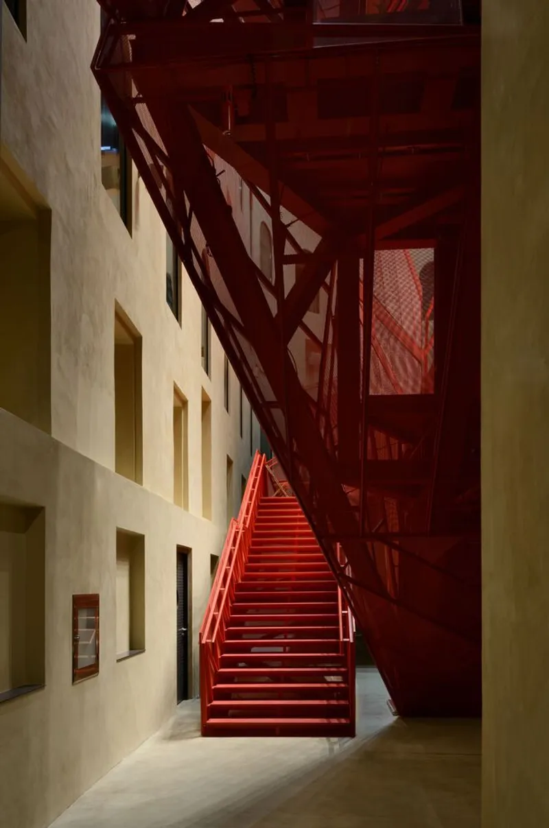 Red steel staircase ascending between plaster wall openings and truss structure under warm artificial lighting