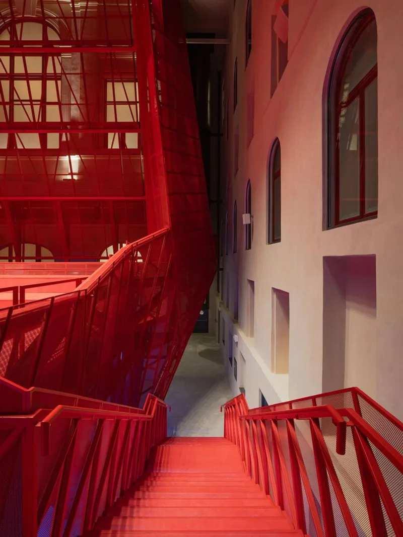 Descending view down red metal staircase between glazed balcony and arched window openings with plaster surround