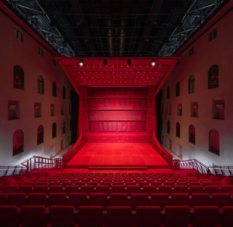 Interior courtyard theatre with red seating stairs and arched wall openings under exposed ceiling trusses