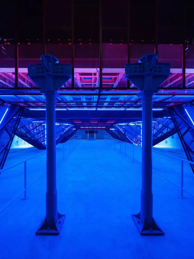 Upward view into exposed steel structural framework illuminated with blue and pink lighting at night