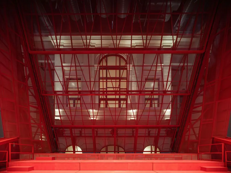 Interior atrium view through red steel scaffolding framework surrounding arched openings with warm lighting