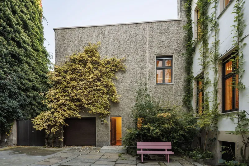 Courtyard view of textured concrete facade with garage door and pink bench flanked by climbing vines