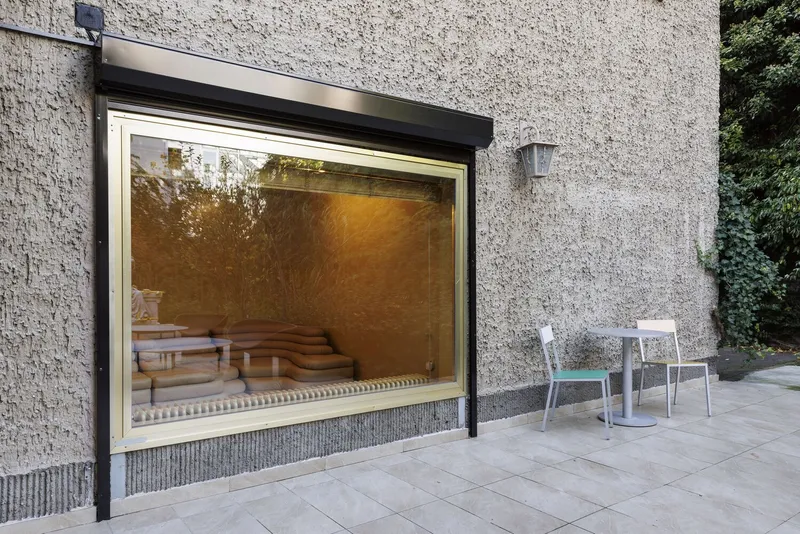 Large window opening in textured concrete wall framing interior seating visible from paved terrace