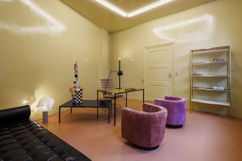 Living area with purple barrel chairs, black desk, open shelving, and cream walls under recessed ceiling cove