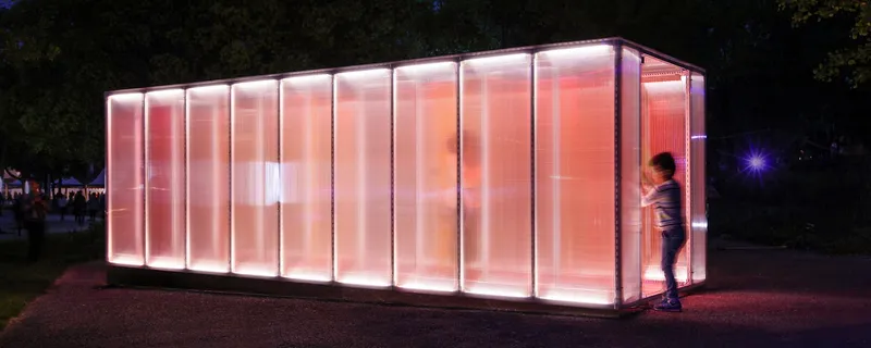 Exterior view of the backlit translucent panel pavilion glowing pink and orange at night with a child running