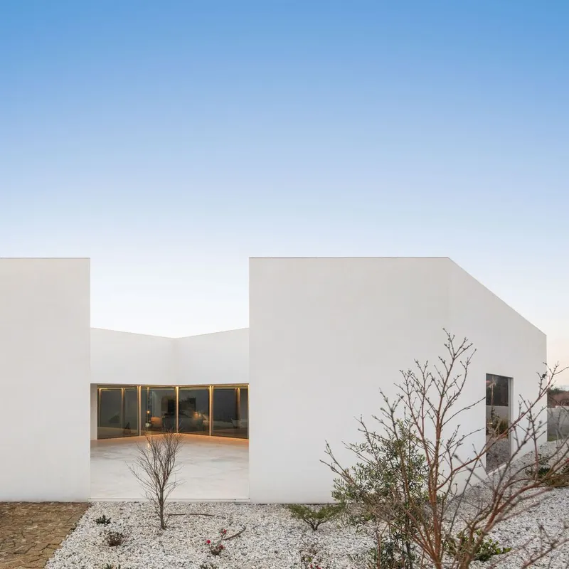 White stucco courtyard with glass doors and young trees planted in white gravel at dusk