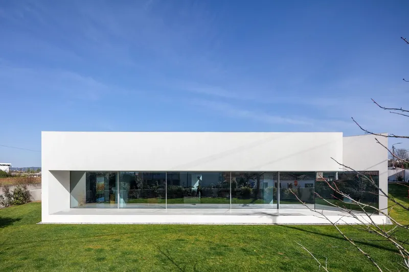 Long white facade with continuous glazing below a hovering roof plane across green lawn