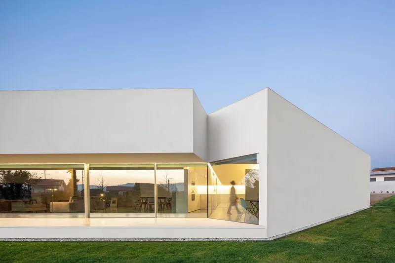 Corner view of the glazed living volume with lawn and a figure passing at twilight