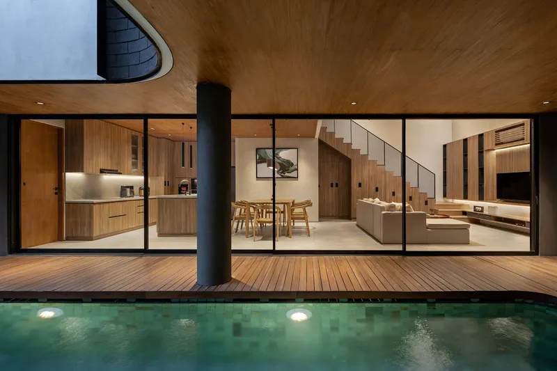 Poolside view of the timber deck and floor-to-ceiling glazing at night with interior lighting