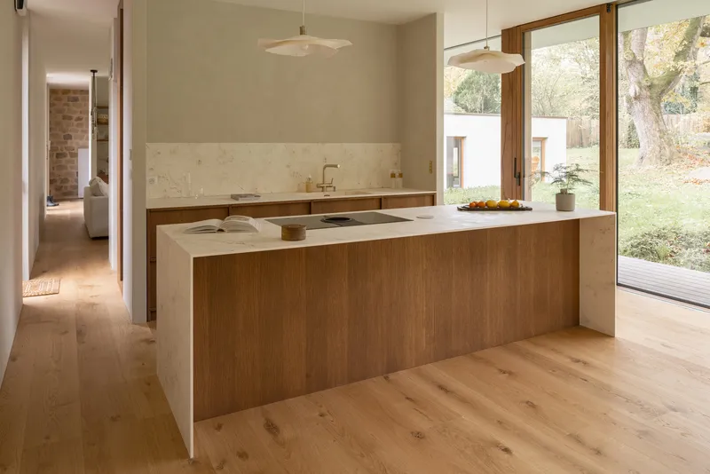 Kitchen island combining stone and timber cabinetry with full-height glazing overlooking garden in autumn