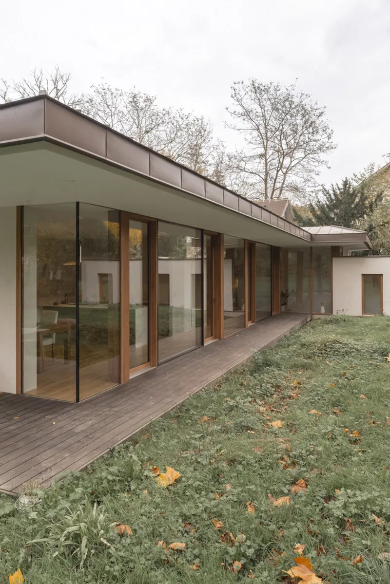 Timber-framed glazed elevation with timber deck terrace extending along sloped lawn scattered with fallen leaves