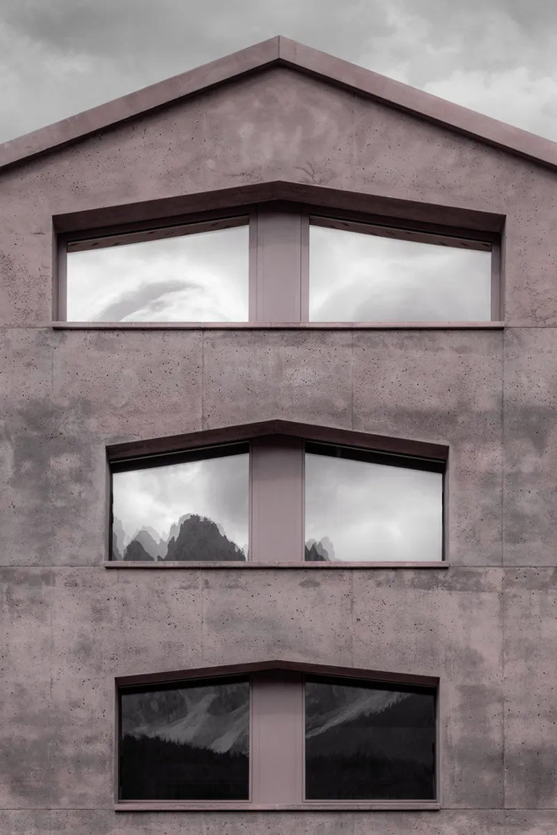 Concrete facade with three stacked corner windows reflecting cloudy skies and mountain trees