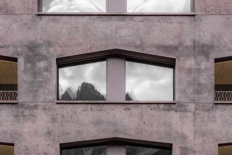 Corner window set in pinkish concrete facade reflecting mountain peaks in the glass panes