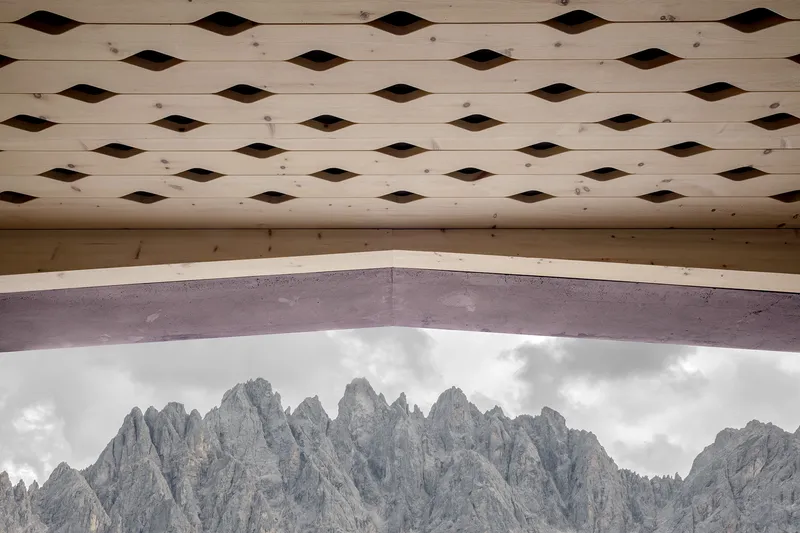 Timber soffit with diamond-shaped apertures framing a view of jagged mountain peaks and clouds