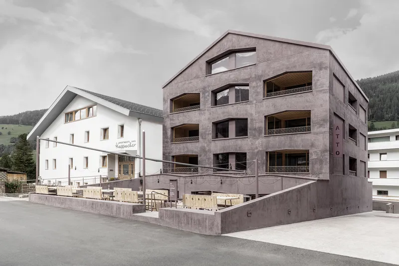 Street view of the concrete residential building with balconies beside a white gabled structure under cloudy skies