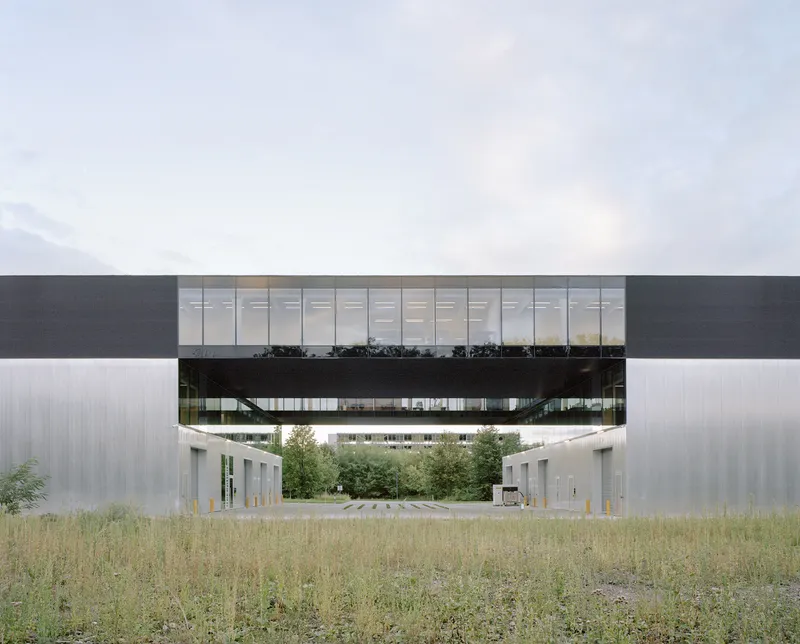Elevated glass bridge spanning between two metal-clad volumes above a gravel field and young trees