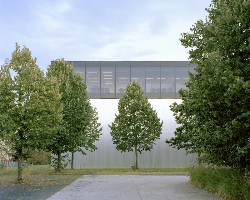Corrugated metal facade with elevated glazed bridge volume framed by mature trees along a gravel path