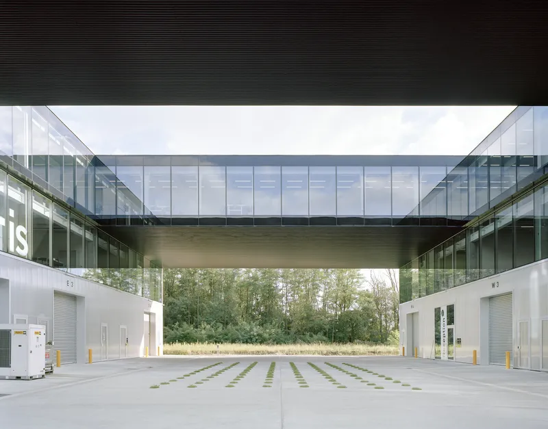 View beneath the glazed bridge connecting two flanking volumes above a paved courtyard with grid pattern