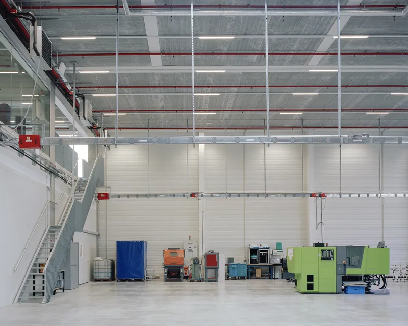Double-height workshop space with mezzanine stair, exposed ceiling infrastructure, and scattered equipment on the floor