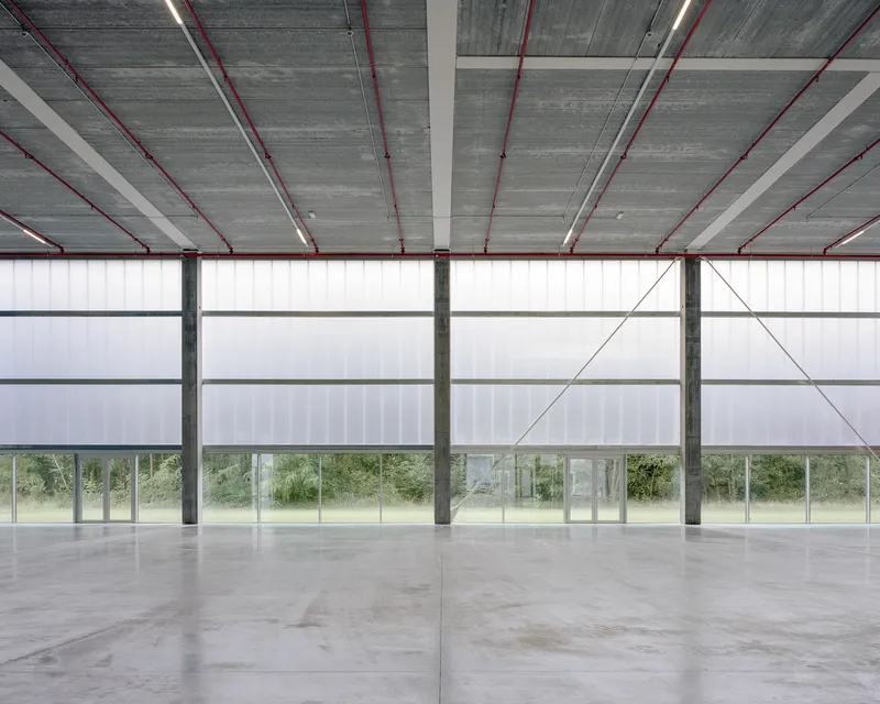 Interior hall with exposed concrete ceiling, red conduits, steel columns, and translucent panel facade overlooking grass