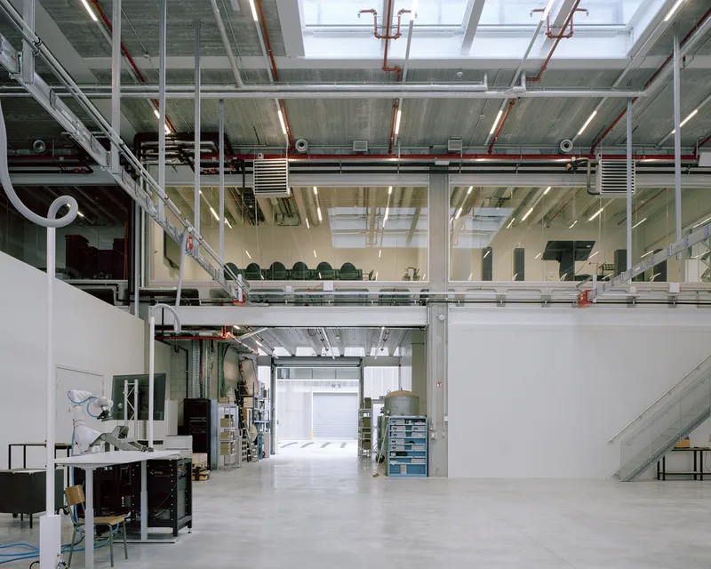 View through mezzanine levels showing exposed concrete slabs, red piping, and workbenches below skylights