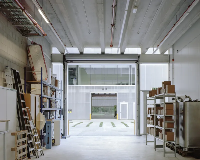 Interior view of workshop space with exposed concrete beams, roll-up door, and material shelving under skylights