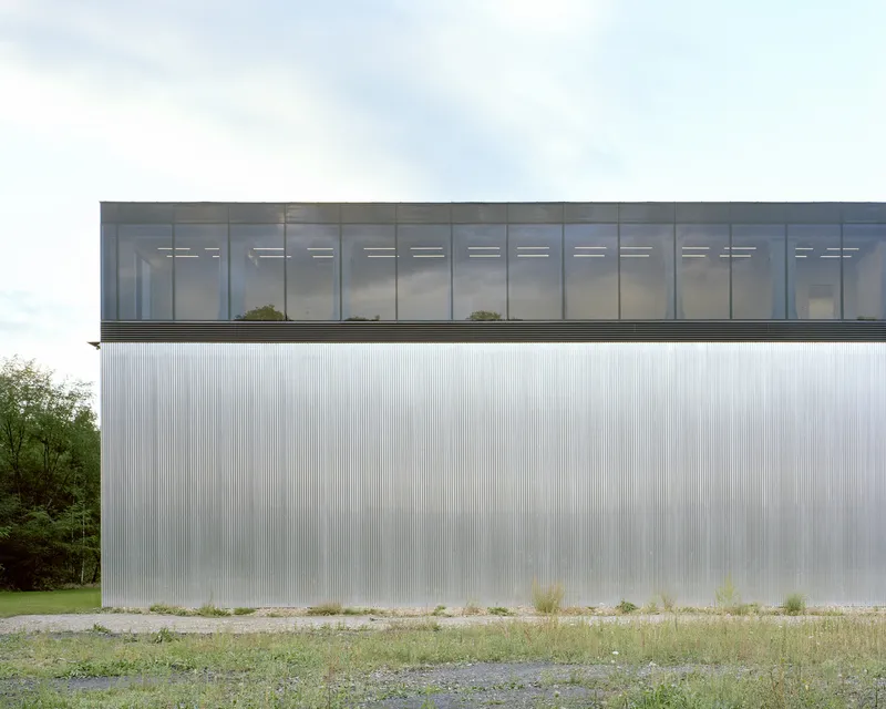 Elevated glass volume above a ribbed metal facade with wildflowers growing at the base