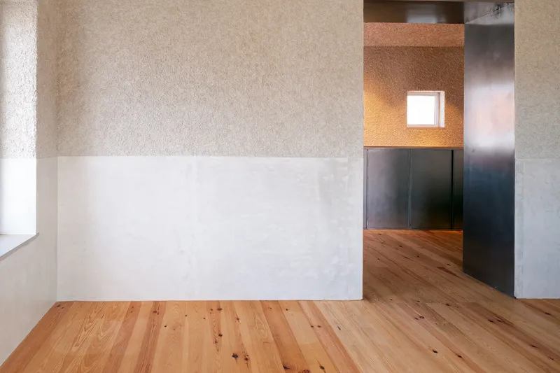 Pine floorboards meeting a plastered partial wall with a backlit window niche beyond dark steel panels