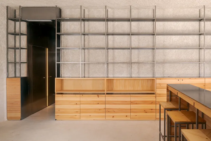 Steel shelving grid behind blonde wood cabinetry and a dark steel doorway on polished concrete flooring