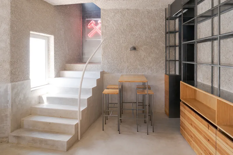Concrete staircase with curved steel handrail ascending past a high table and timber cabinetry below a neon sign