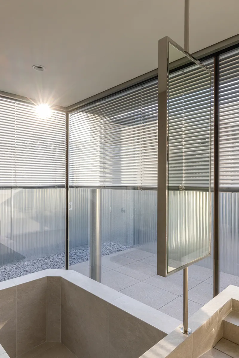 Sunken bath beside floor-to-ceiling louvered glass panels overlooking a gravel courtyard at sunset