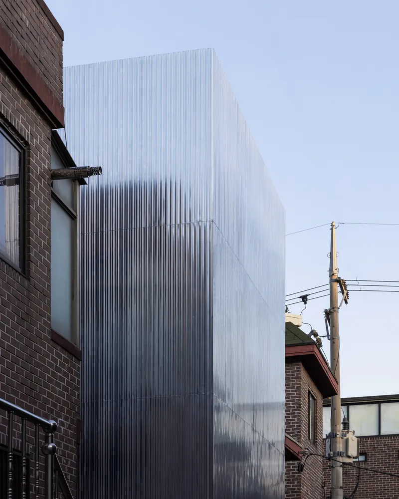 Translucent ribbed metal facade rising between brick buildings and a utility pole at dusk